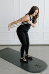 Woman exercising with grey light resistance band on a mat in a room with white paneled walls.